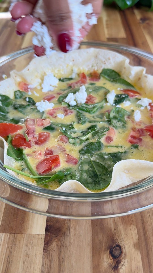 Hand sprinkling feta over eggs with spinach and tomatoes in a glass dish on a wooden table.