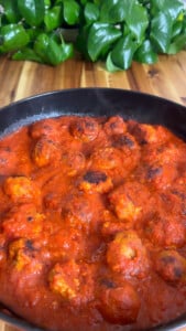 Meatballs simmering in rich tomato sauce in a black pan with fresh greenery in the background on a wooden table.