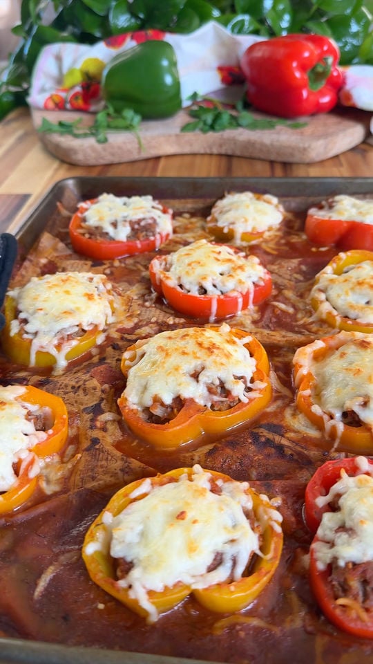 Baked stuffed bell peppers with melted cheese on a tray, fresh peppers and herbs in the background.