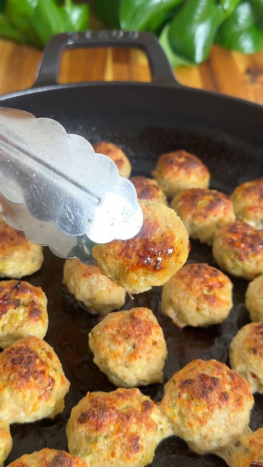 Tongs holding a cooked meatball above a pan filled with browned meatballs, green leaves in background.