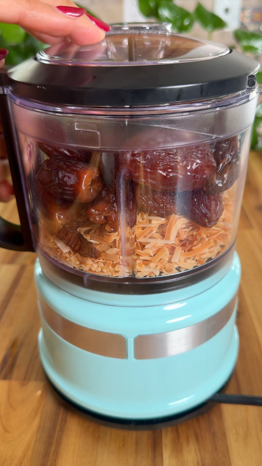Food processor with dates and shredded coconut on a wooden surface.