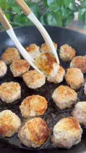 Cooking pan filled with golden-brown meatballs, some being lifted with tongs.