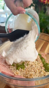 Mixing ricotta, breadcrumbs, and pesto with meat in a glass bowl.