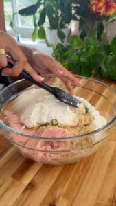 Mixing ground chicken, ricotta, and breadcrumbs in a glass bowl on a wooden countertop.