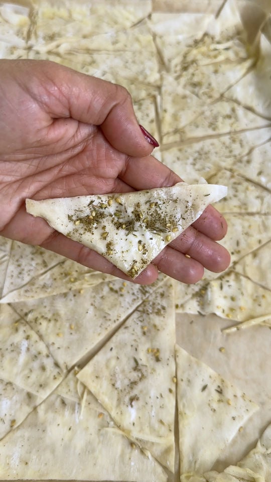 Hand holding a triangular pastry piece with herbs, part of a sheet of similar pastries in the background.