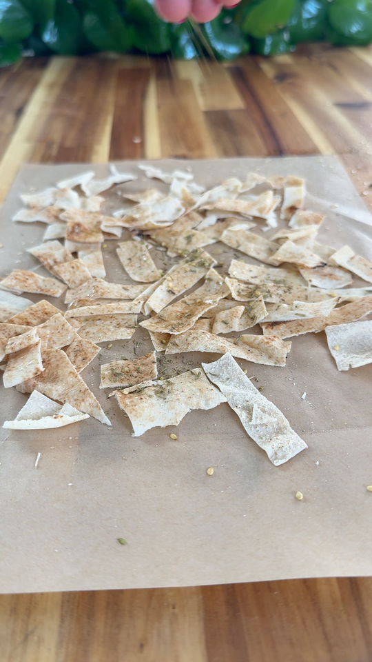 Pieces of pita seasoned with herbs scattered on parchment paper on a wooden table.