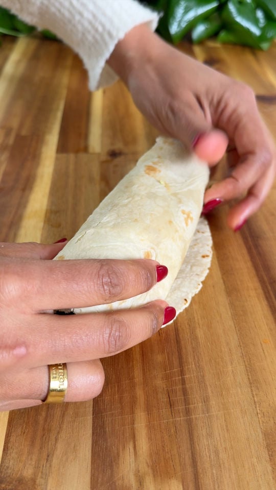 Hands rolling a wrap on a wooden surface with a green plant in the background.