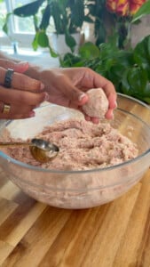 Person forming meatballs from ground chicken mix in a glass bowl on a wooden table.