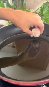 Hand placing meatball into a black pan with oil for cooking.