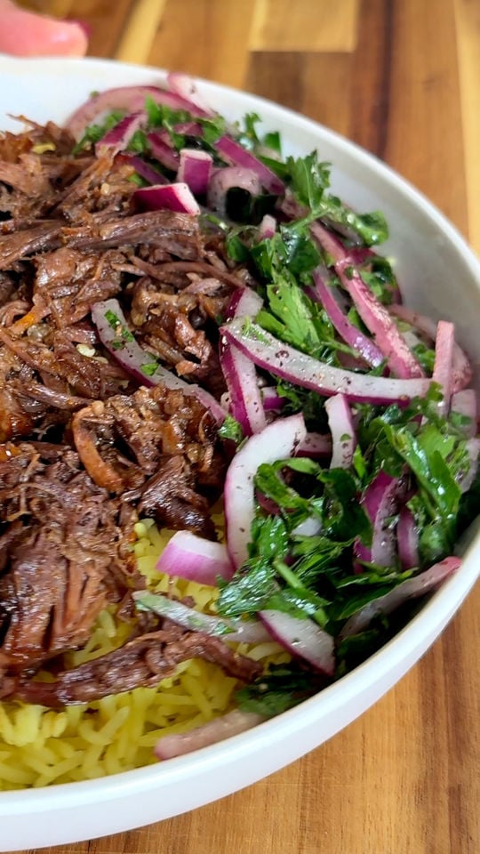 Bowl of shredded beef, yellow rice, and fresh salad with red onions and herbs on a wooden table.