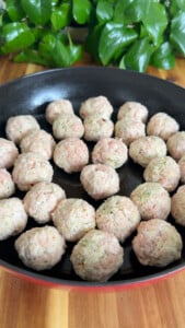 Raw meatballs in black skillet on wooden table with green plants in background.