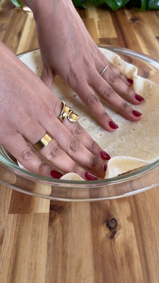 Hands placing tortilla in glass dish on wooden table.