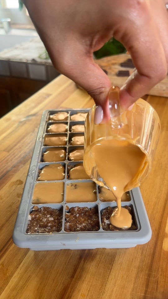 Pouring almond butter into ice cube tray filled with crumbled mixture on wooden surface.