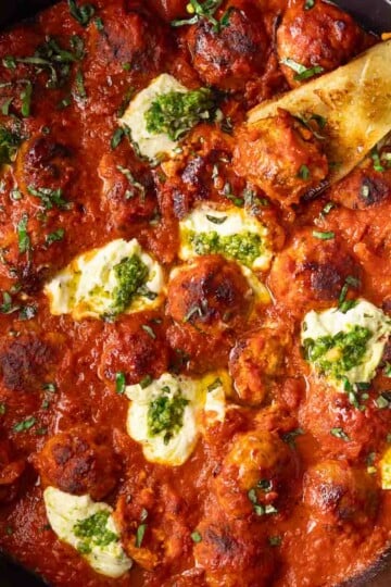 Ricotta meatballs in tomato sauce with dollops of cheese and pesto, garnished with fresh basil in a black skillet.