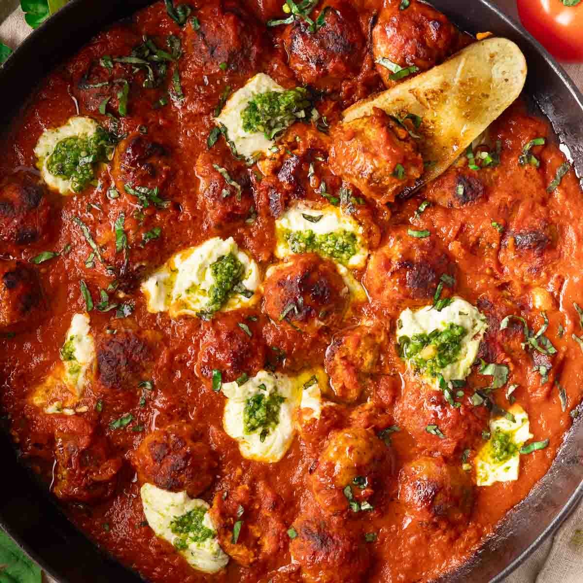 Ricotta meatballs in tomato sauce with dollops of cheese and pesto, garnished with fresh basil in a black skillet.
