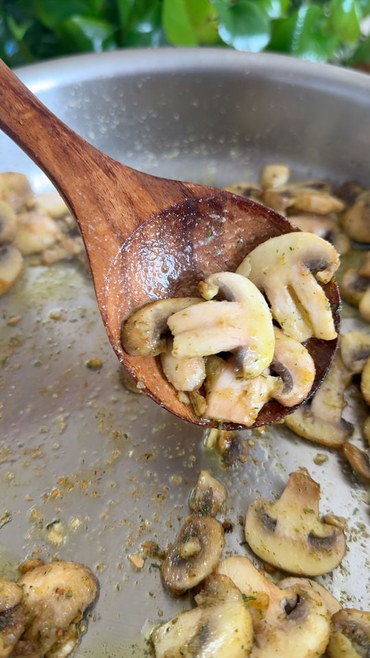 Sautéed mushrooms on a wooden spoon over a pan with herbs and seasoning visible.