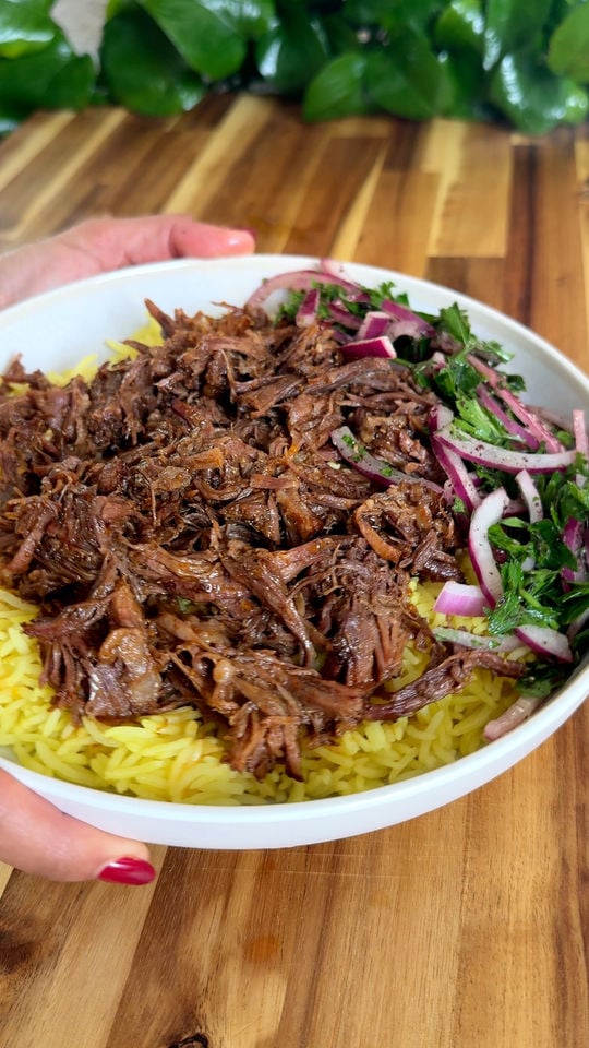 Shredded beef over yellow rice with red onions and cilantro salad in a white bowl on a wooden table.