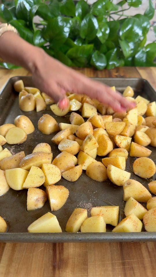 Hand seasoning sliced potatoes on a baking tray with green plant in the background.