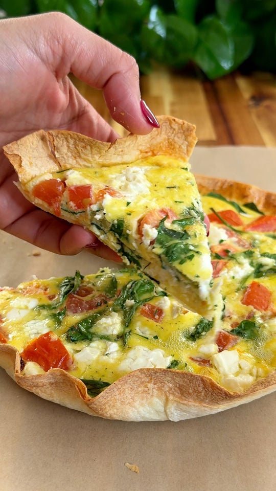 Hand holding a slice of egg tortilla bake with tomatoes, spinach, and cheese on a wooden background.