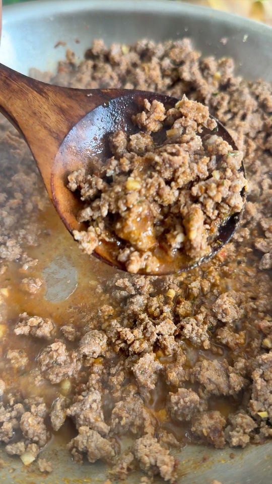 Wooden spoon stirring cooked ground beef in a pan with visible spices and water.