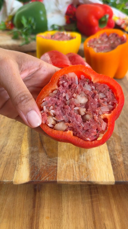 Hand holding a red bell pepper stuffed with minced meat and onions, with more stuffed peppers in the background.