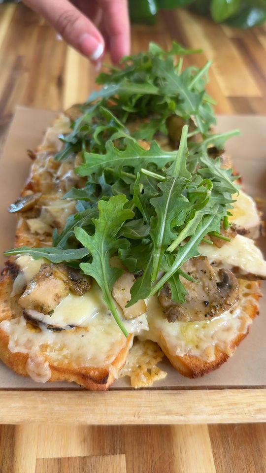 Toasted bread topped with melted cheese, mushrooms, arugula, on wooden board.
