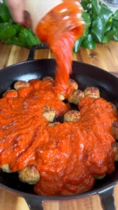 Person pouring tomato sauce over meatballs in a skillet, with green plant in the background.