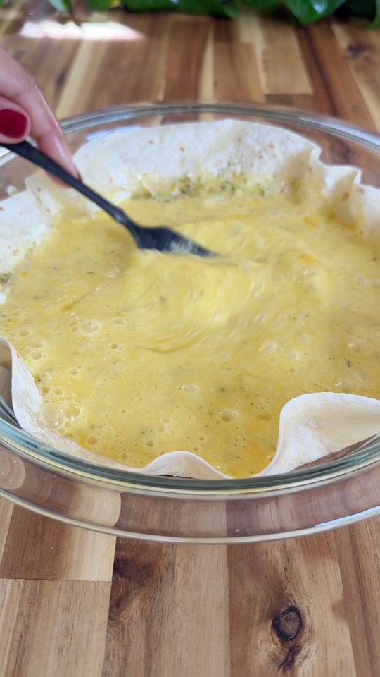 Mixing eggs in a tortilla-lined glass bowl with a fork on a wooden surface.
