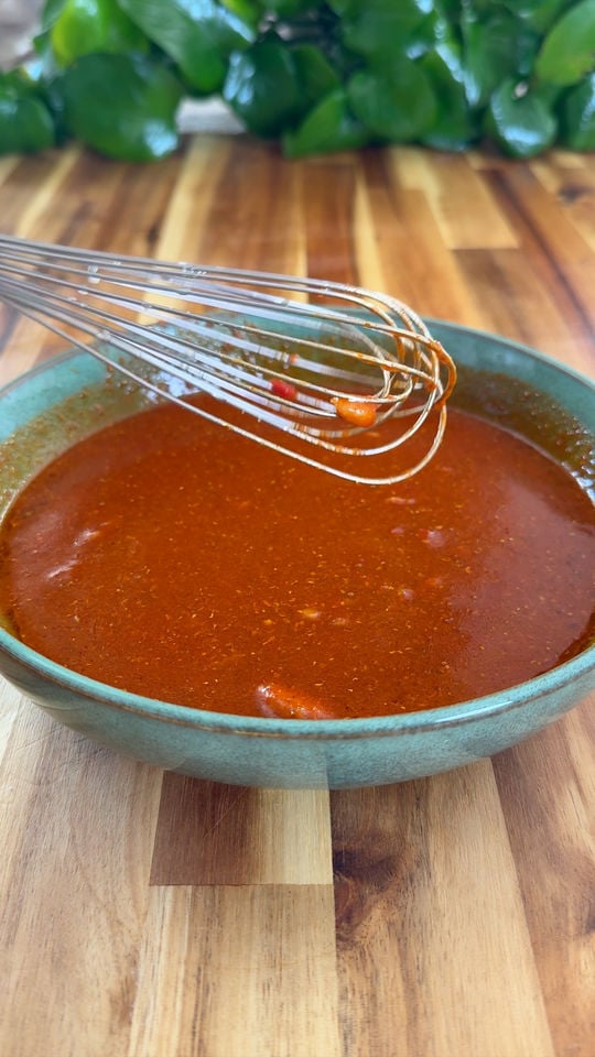 Green bowl with red sauce and a whisk, on wooden table, green plant in background.