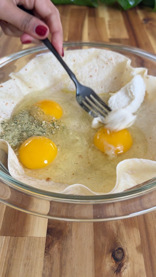 Hand whisking eggs, spices, and Greek yogurt in a tortilla-lined glass bowl on a wooden surface.