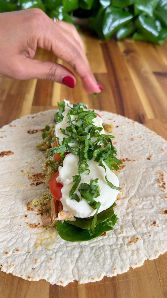 Hand adding basil to a wrap filled with spinach, mozzarella, tomato, and tuna on a wooden table.