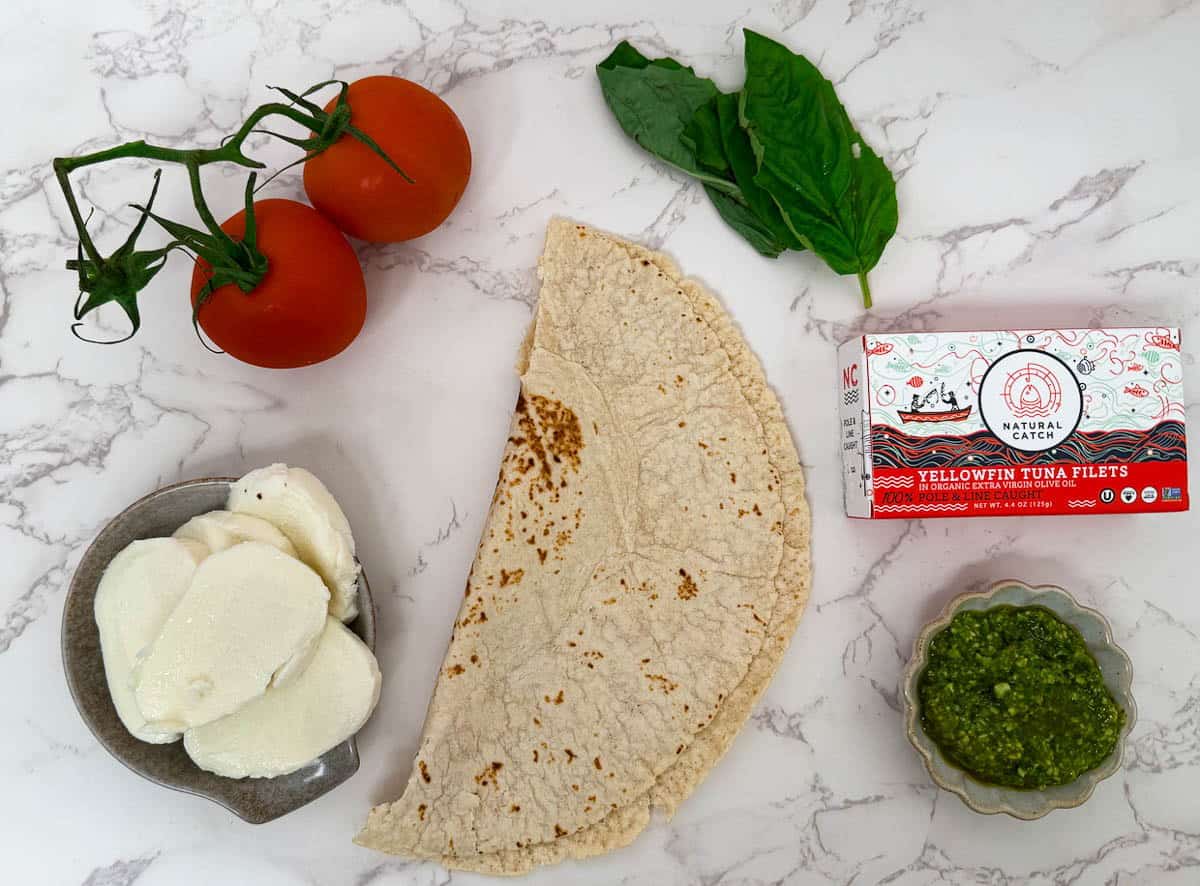 Tortilla, tomatoes, tuna fillets, basil, pesto, and mozzarella on a marble surface.