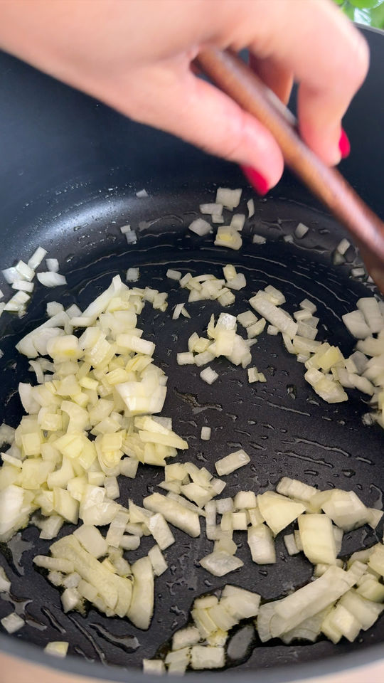 Chopped onions sautéing in a black pan, stirred by a hand holding a wooden spoon.