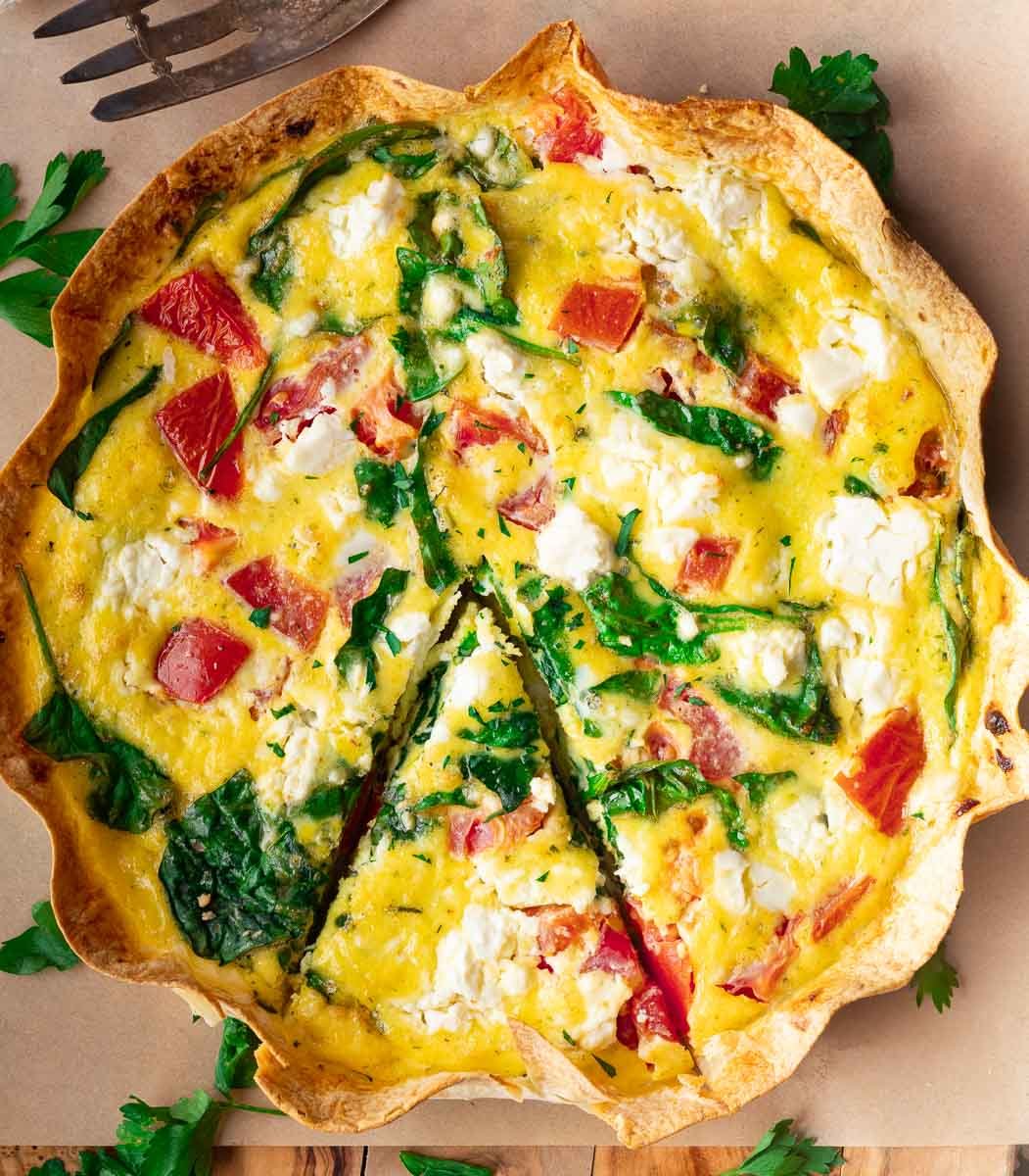 Egg tortilla bake with spinach, tomatoes, and cheese on a wooden table, one slice cut out.