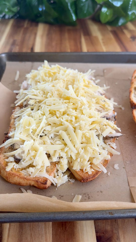 Toasted bread topped with grated cheese on a parchment-lined baking tray, wooden table background.