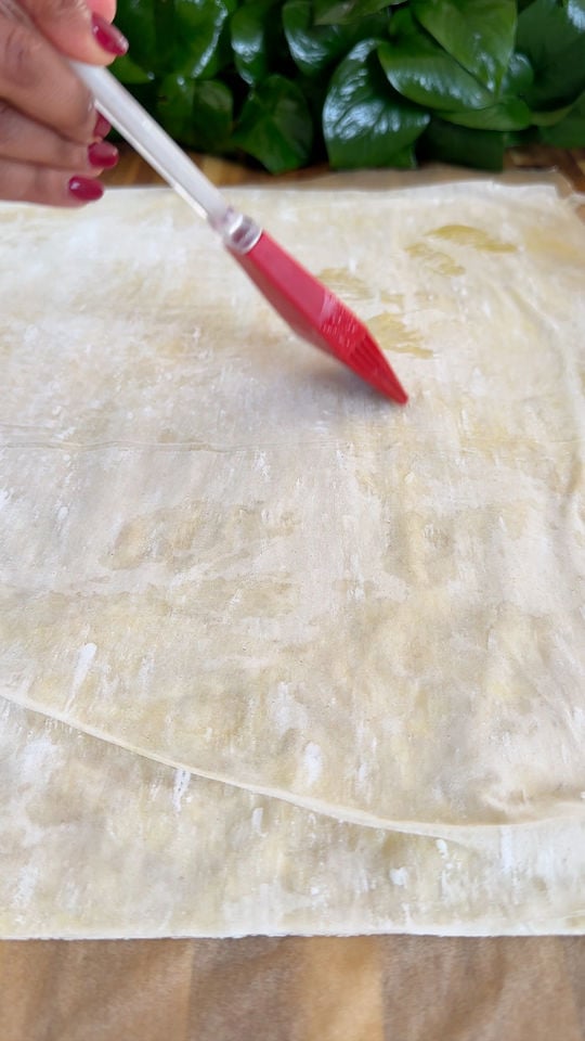 Hand brushing oil on a sheet of pastry dough with a red silicone brush, plants in the background.
