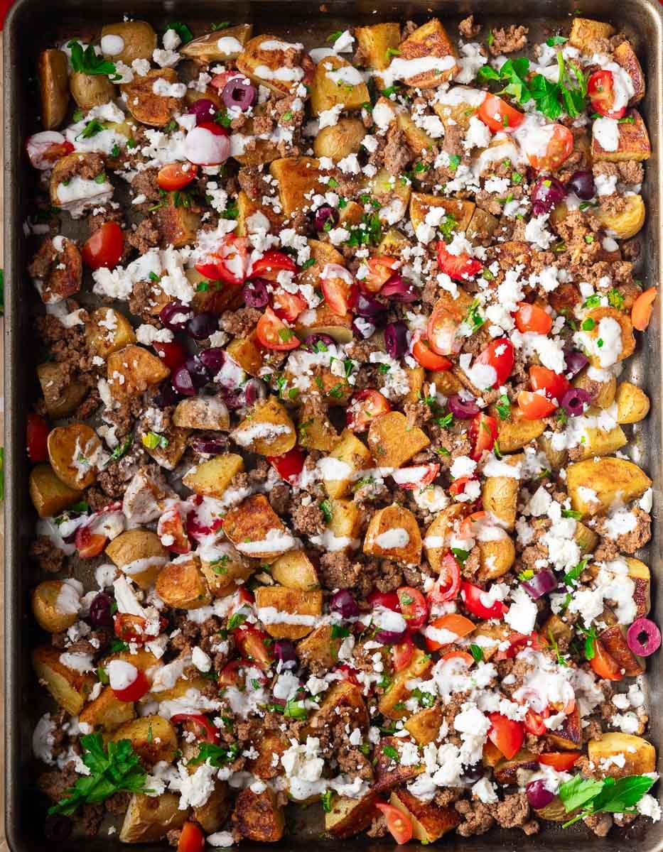 mediterranean ground beef potato bowl with roasted potatoes, ground meat, tomatoes, olives, feta cheese, and herbs on a baking sheet with a creamy sauce.