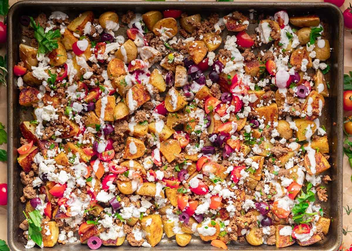Roasted potatoes with ground meat, cherry tomatoes, olives, feta cheese, and herbs on a baking tray.