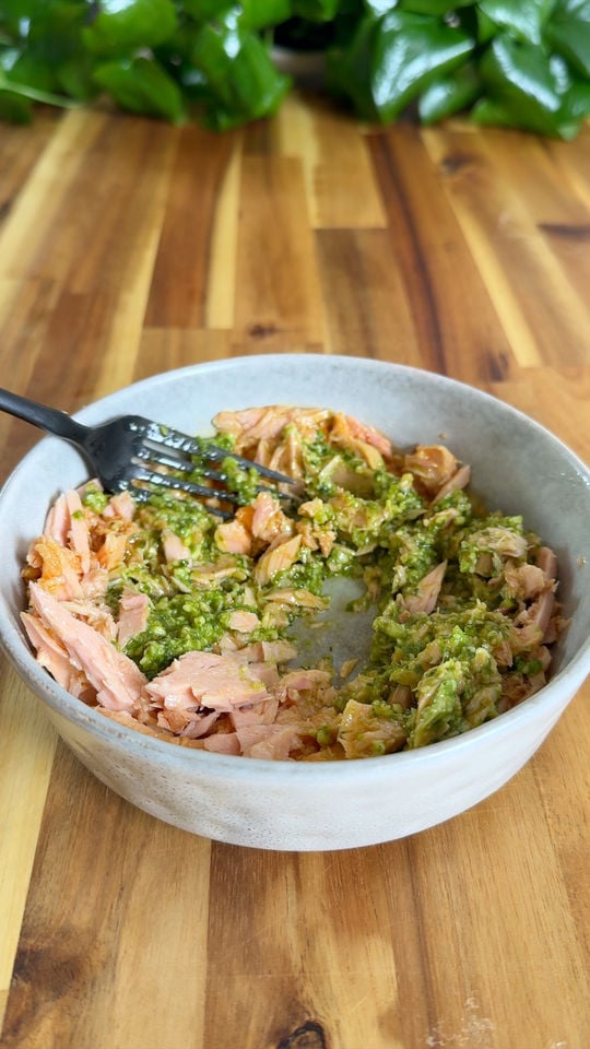 Bowl of shredded tuna mixed with green pesto sauce and black fork on wooden table background.