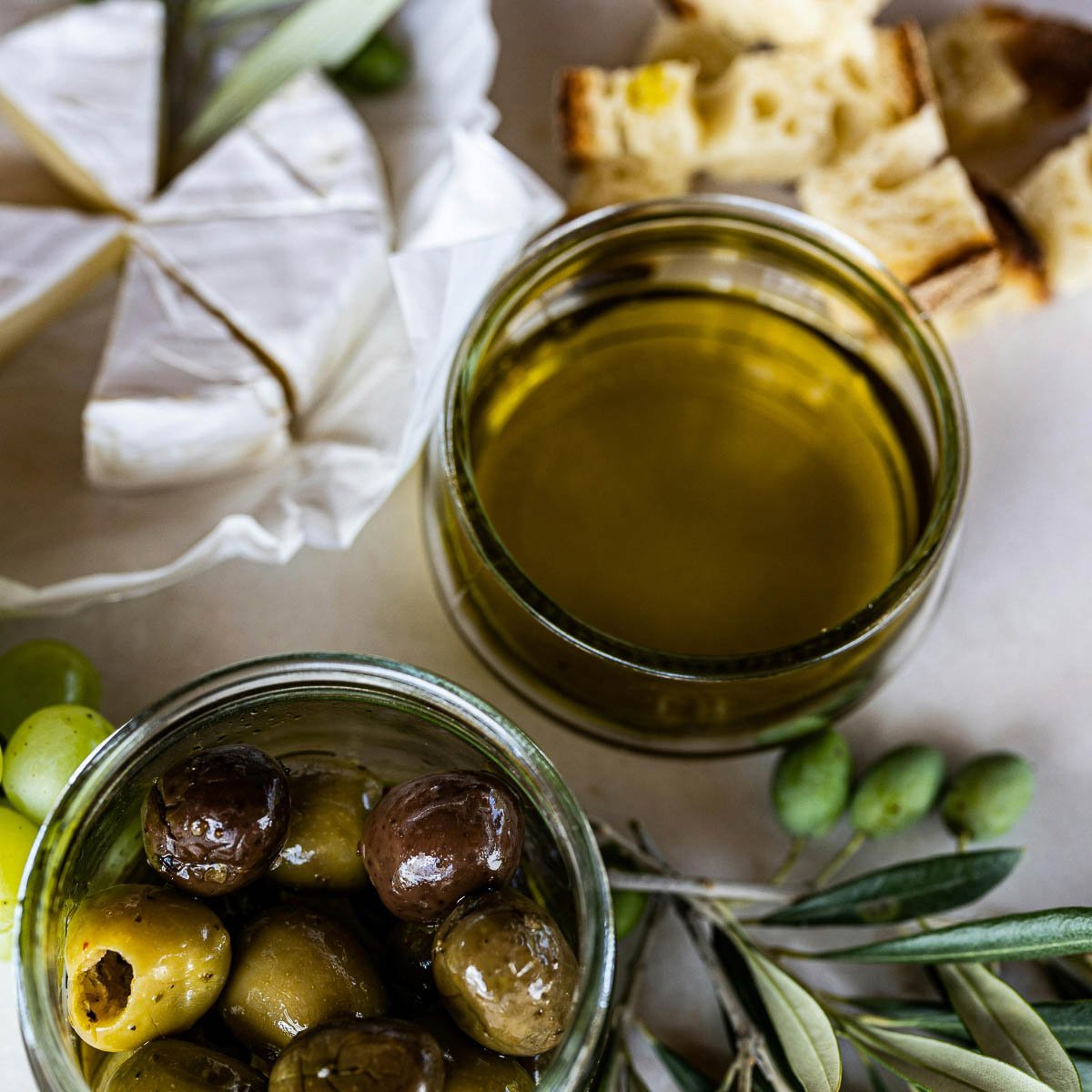Olives in a jar, olive oil in a glass, camembert cheese, and sliced bread on a light background.