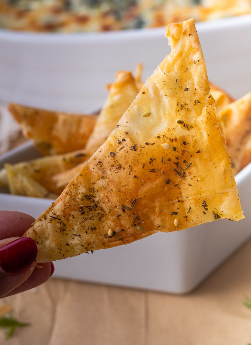 Close-up of a seasoned Phyllo chip held by hand, with more chips in a white bowl in the background.