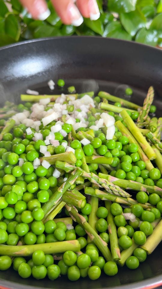 Fresh asparagus, peas, and chopped onions cooking in a black pan with a hand above.