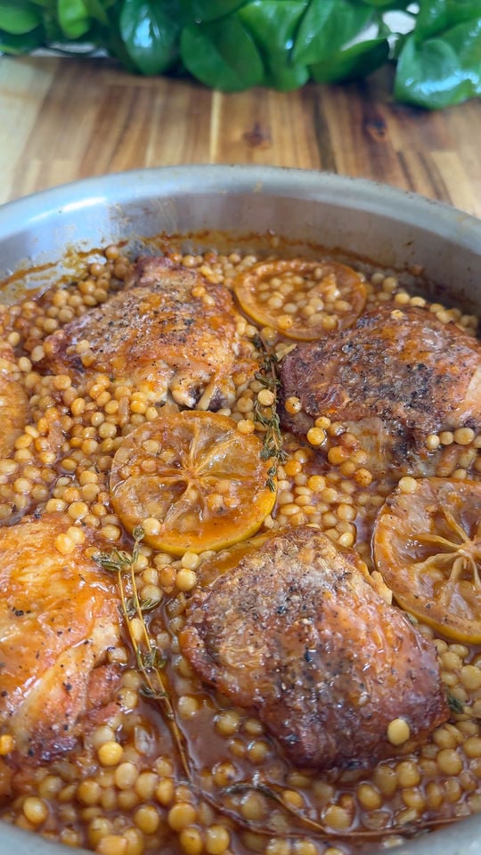 Baked chicken thighs with lemon slices, pearl couscous, and herbs in a round pan on a wooden table.