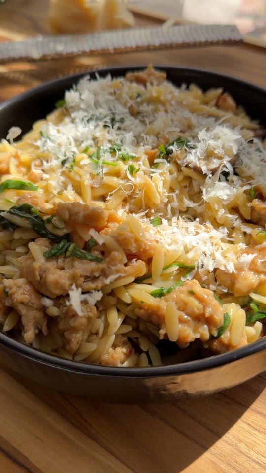 Bowl of orzo pasta with spinach, shredded cheese, and sausage on a wooden surface.