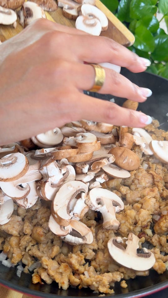 Hand adding sliced mushrooms to a pan with cooked sausage.