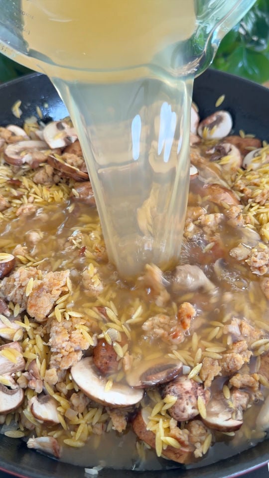 Pouring broth into a pan with orzo, mushrooms, and sausage.