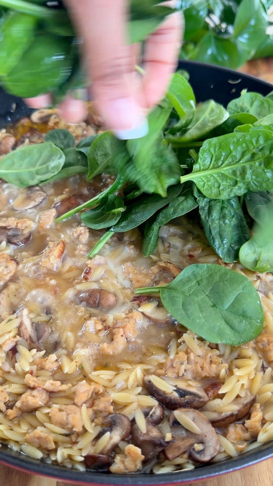 Hand adding fresh spinach to a pan of orzo pasta with mushrooms and crumbled sausage.