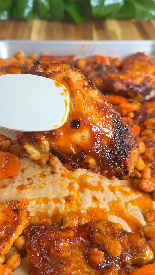 Grilled chicken with glaze held by tongs over a tray of roasted chickpeas and carrots.