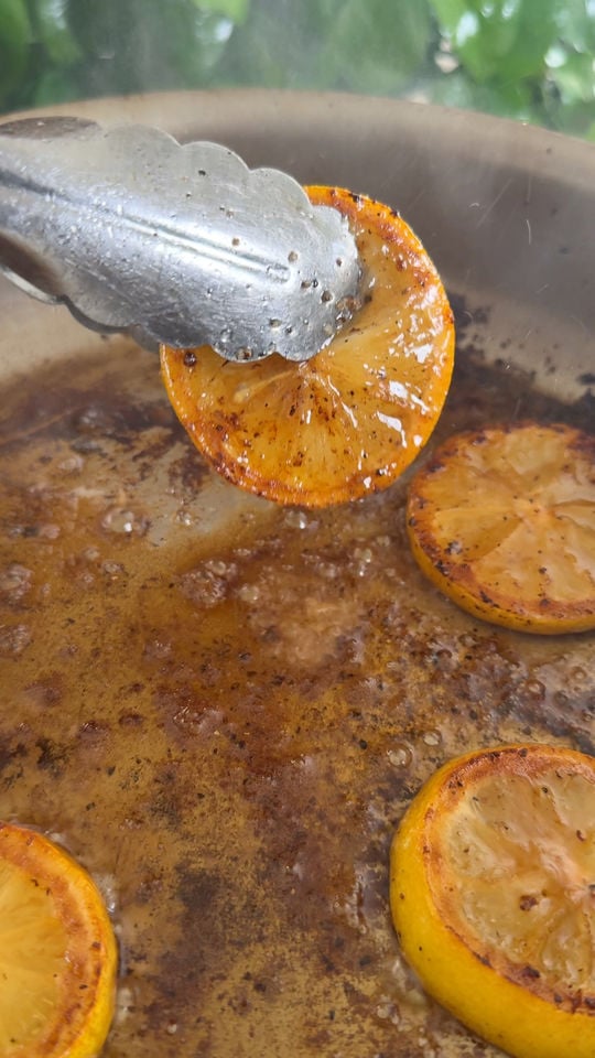 Tongs holding grilled lemon slice over sizzling pan with browned lemon pieces.