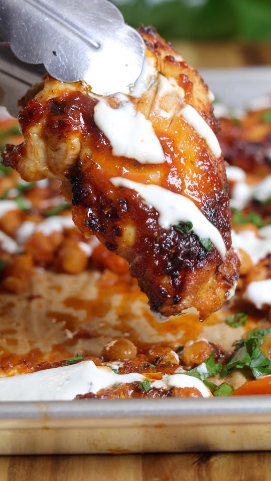 Honey harissa chicken tray bake with sauce, topped with creamy dressing, held by tongs over roasted chickpeas on a baking tray.